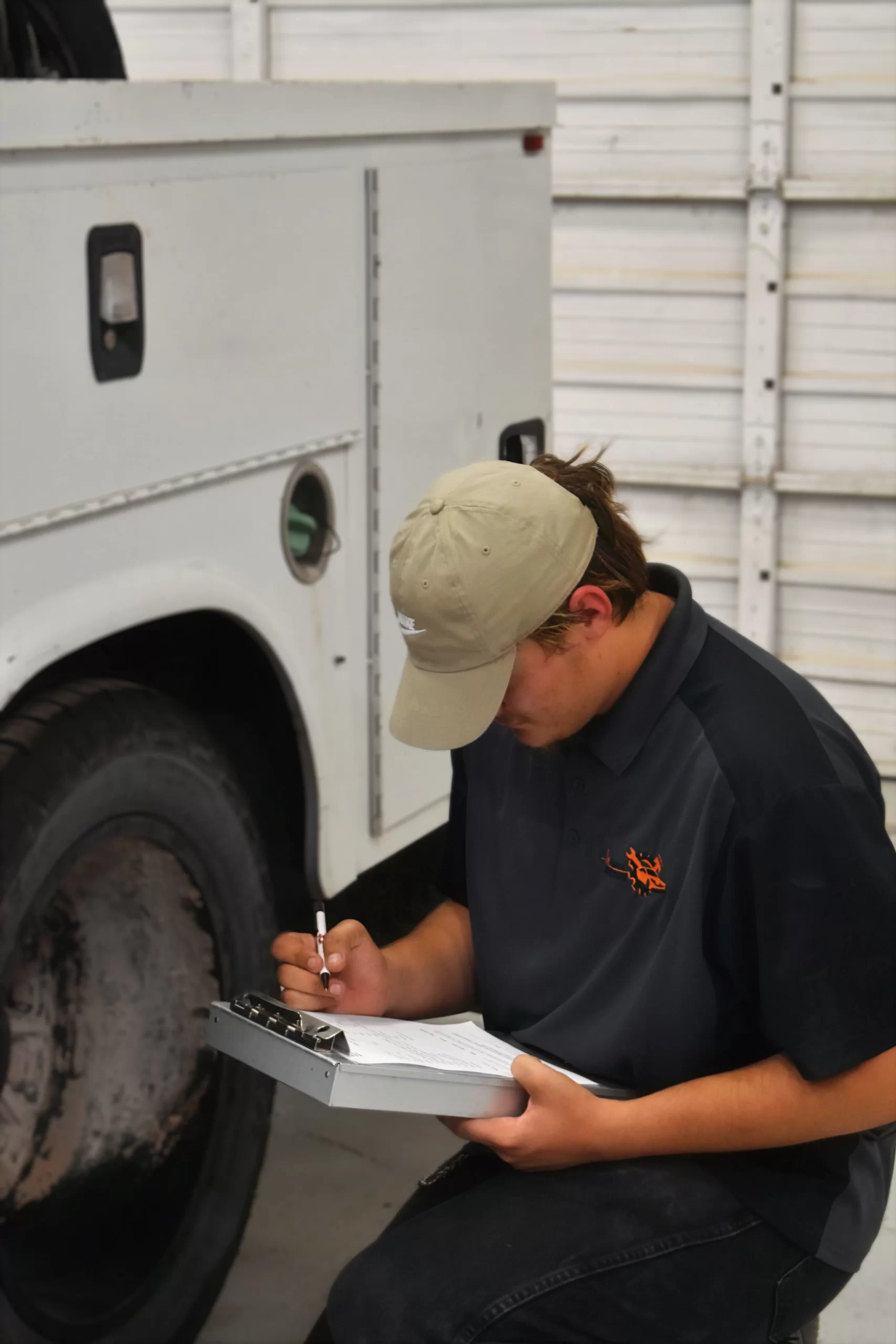 Mechanic kneeling by a truck, filling out paperwork on a clipboard in a garage setting.