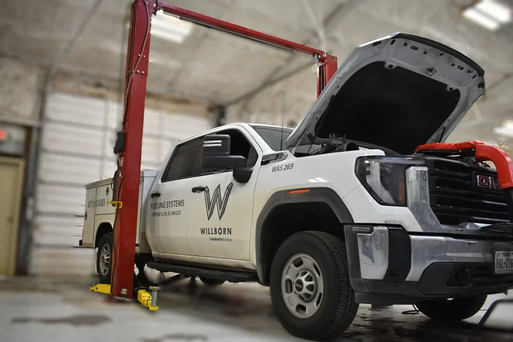 White GMC service truck on a red lift in a garage, hood open for maintenance, labeled "Willborn."