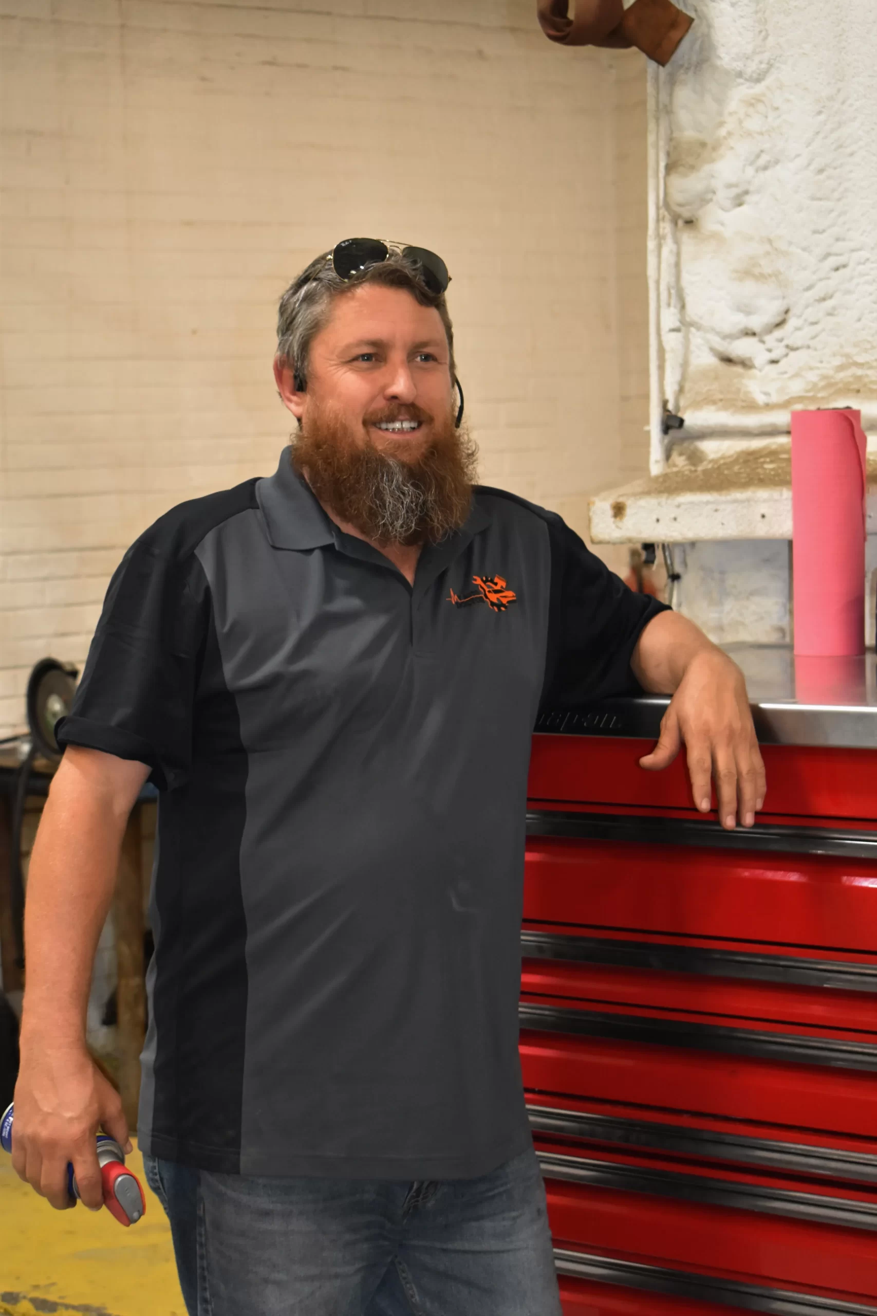 Smiling mechanic leaning against a red tool chest, holding a tool, in a workshop.